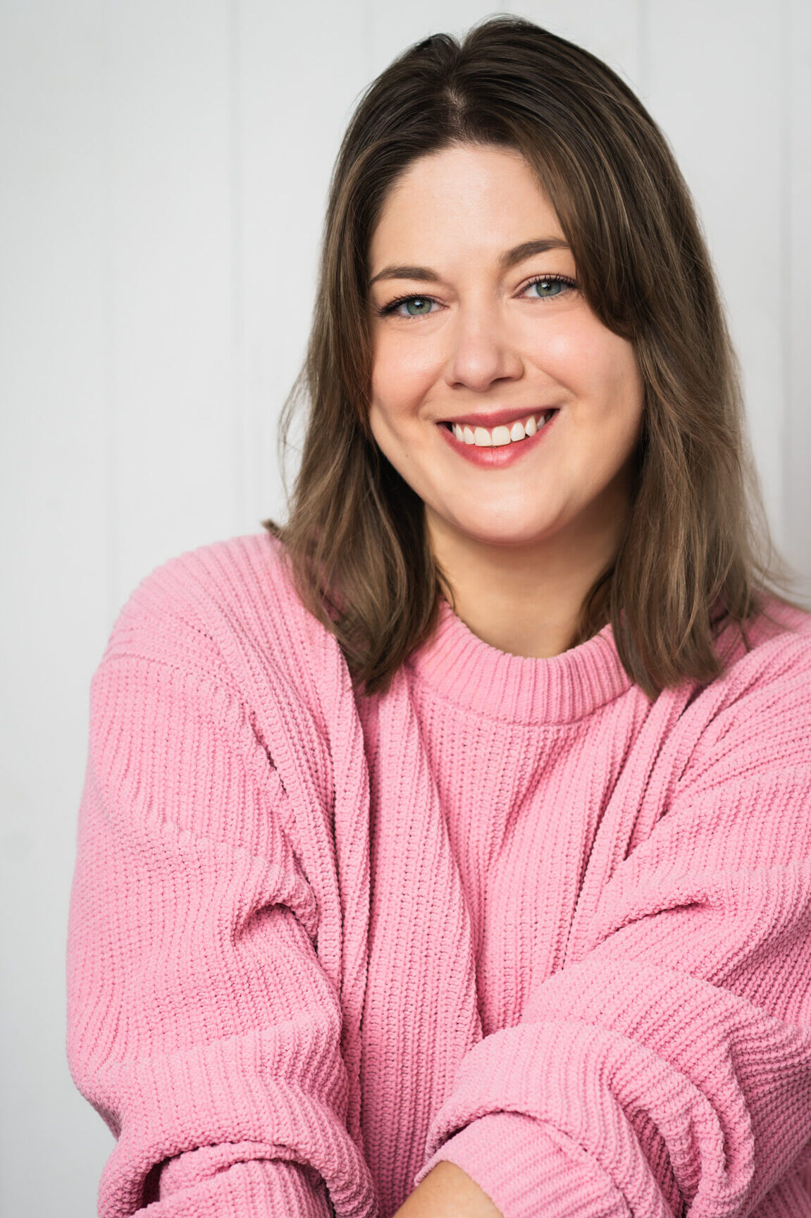 White woman with light brown hair smiles. Hands resting on her knees. She is wearing a pink sweater and light blue jeans.
